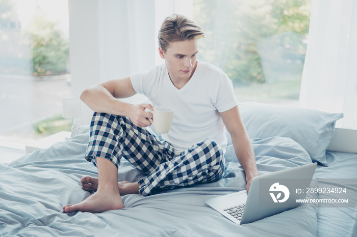 Full size photo of concentrated blonde hair man use his laptop for job make project his boss want hold mug with coffee sit on bed wear checkered plaid pajama in house indoors