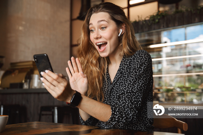 Emotional happy beautiful young pretty woman sitting in cafe indoors using mobile phone talking with friends take a selfie.