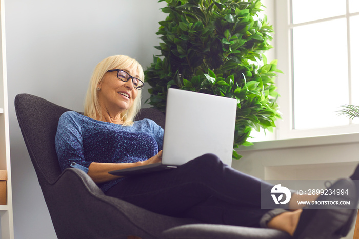 Mature woman with glasses sitting in an easy chair and working at home using a laptop. Senior woman doing business remotely sitting in a comfortable home office. Online business concept.