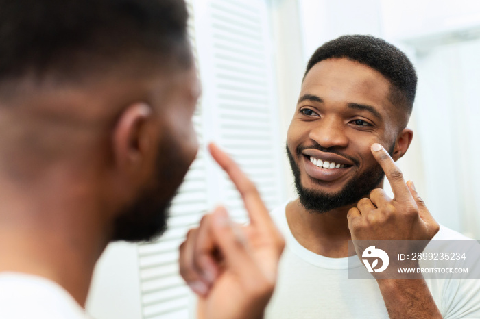 Cheerful black man applying pampering cream on face in bathroom