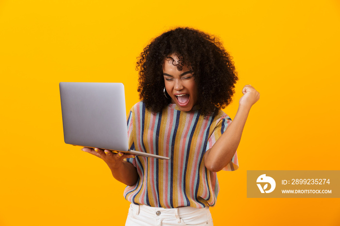 Emotional african woman posing isolated over yellow background using laptop computer make winner gesture.