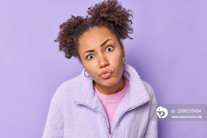 Curly haired beautiful young woman looks with attentive gaze at camera pouts lips focused at something dressed in warm fur jacket listens information attentively isolated over purple background.