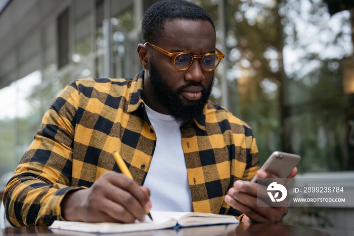 Pensive African American man using mobile phone, taking notes, working freelance project online, sitting outdoors. Serious student studying, learning something