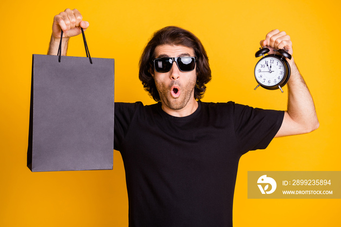 Photo of young man showing alarm clock dial mall packet surprised expression t-shirt sunglass isolated yellow color background