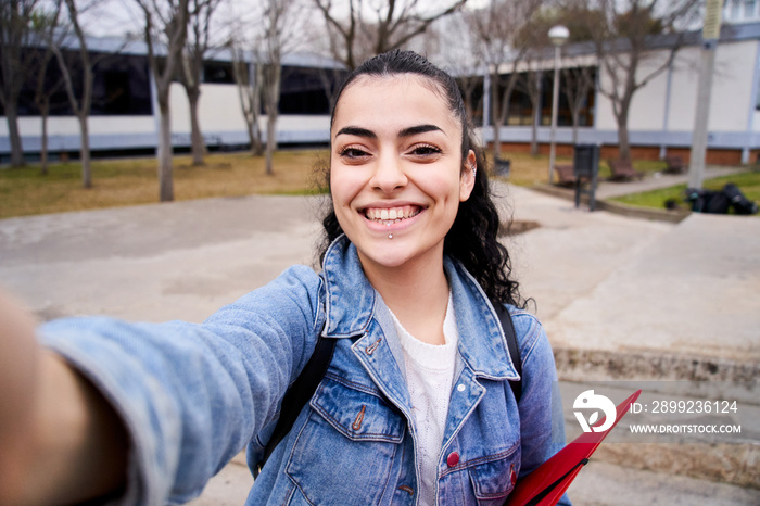 Selfie of a cheerful and happy Caucasian teenager looking smiling at camera in high school playground. Portrait of a young female student on college campus. Women and social media content.