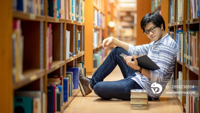 Smart Asian man university student wearing glasses reading book by vintage bookshelf. Textbook resources in college library for educational subject and research. Scholarship for education opportunity.