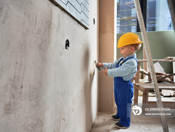 Little boy builder worker measuring wall with construction ruler. Cheerful kid in safety helmet and work overalls working on home renovation.