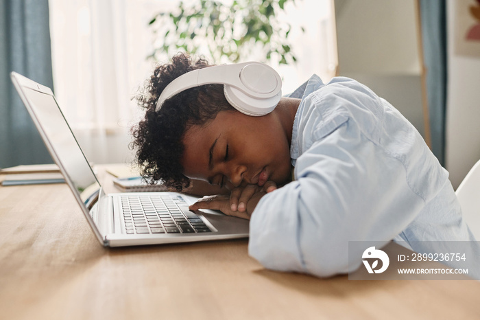 African tired boy in headphones sleeping at his desk with laptop after online lesson at home