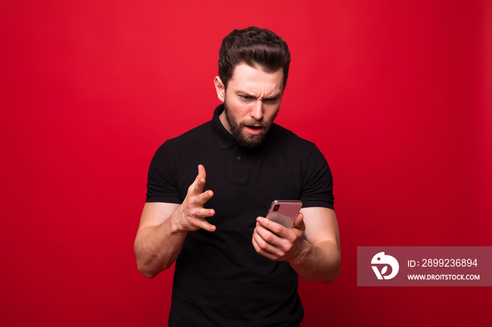 Portrait of screaming angry young bearded emotional man standing over red wall background isolated. Looking aside while talking by phone.