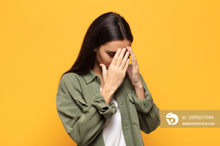 young hispanic woman covering eyes with hands with a sad, frustrated look of despair, crying, side view