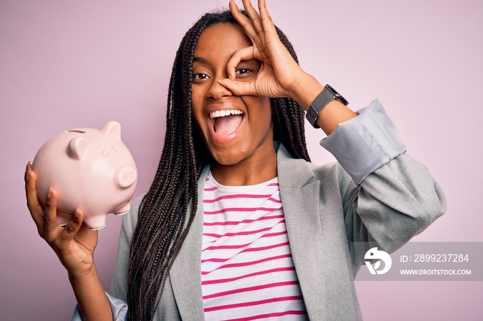 Young african american business woman saving money on piggy bank over isolated background with happy face smiling doing ok sign with hand on eye looking through fingers