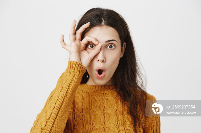 Close-up studio portrait of funny attractive dark-haired woman showing okay sign near eye and looking through it as if it is monocle, expressing surprise, standing over gray background.