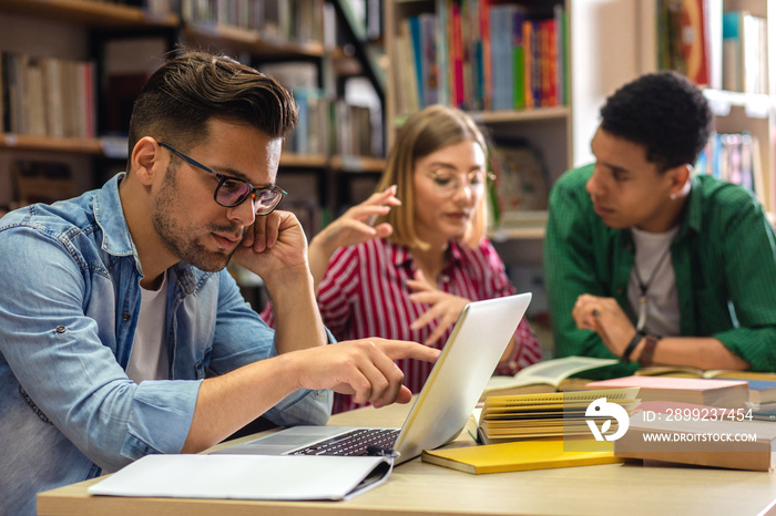 Three young students study in the school library, male student using laptop for researching online.