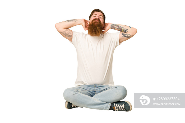 Young adult redhead with a long beard sitting on the floor isolated feeling confident, with hands behind the head.