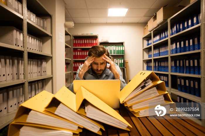 Worried businessman with laptop in file storage room