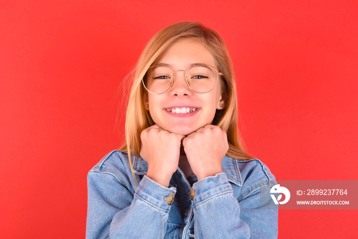 Satisfied blonde little kid girl wearing denim jacket over red background touches chin with both hands, smiles pleasantly, rejoices good day with lover