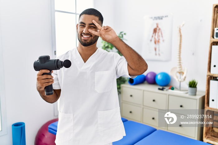 Young indian physiotherapist holding therapy massage gun at wellness center doing peace symbol with fingers over face, smiling cheerful showing victory