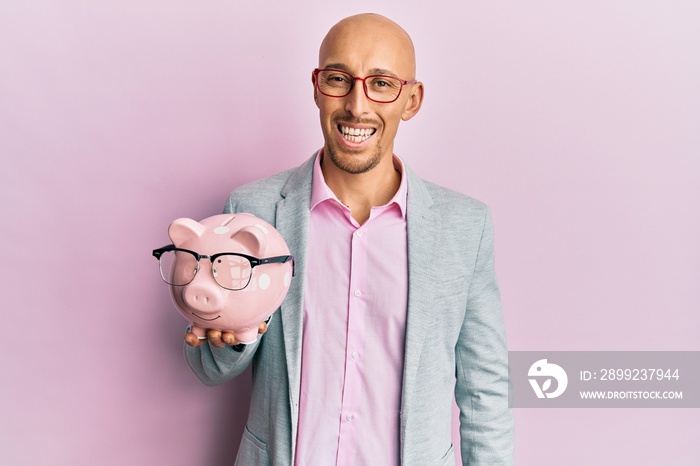 Bald man with beard holding piggy bank with glasses looking positive and happy standing and smiling with a confident smile showing teeth