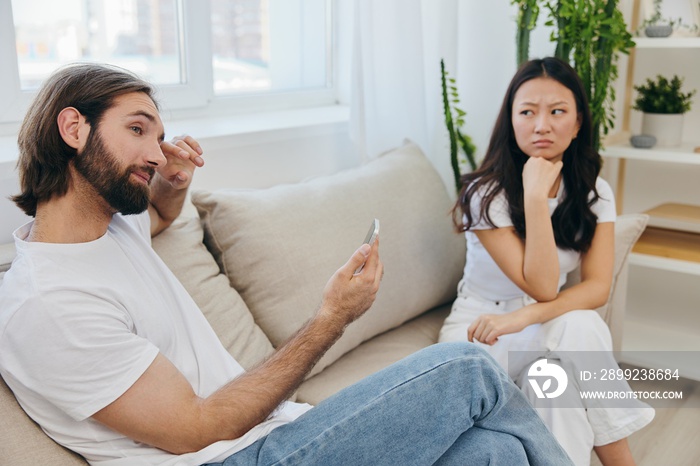 A man looks at the phone screen during an argument with his girlfriend. The angry and hurt woman looks in his direction and is sad. Family discord at home, phone addiction