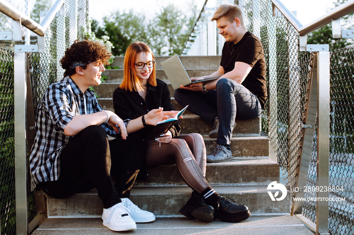 Group of three young attractive college students sitting studying together using laptop in university campus stairs.