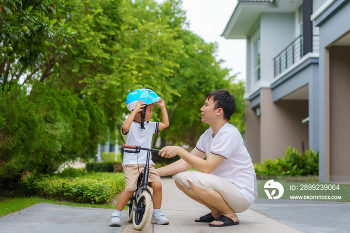 Asian father wears a helmet for his son while teaching his child how to ride a bicycle in a neighborhood garden, fathers interact with their children throughout the day.