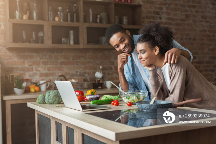 African-american couple cooking dinner with recipe on laptop