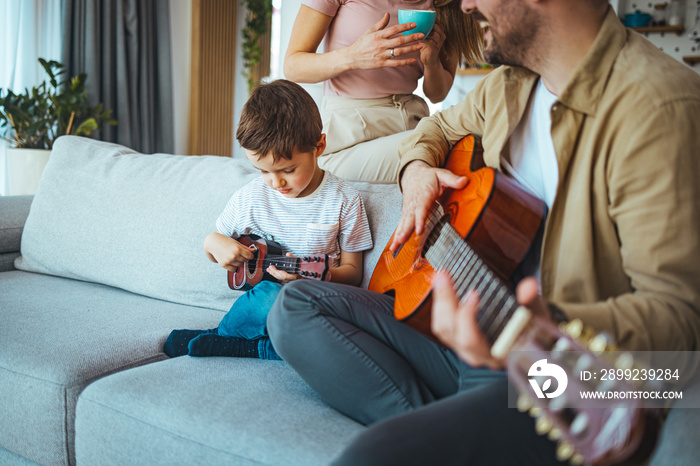 Father teaching his son to play guitar at home. The father teaches his son to play the guitar. He helps him pick up guitar chords. Mom watching. They are in a good mood.