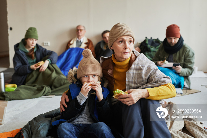 Distressed Caucasian refugee family eating sandwiches while hiding in shelter on floor covered with blankets