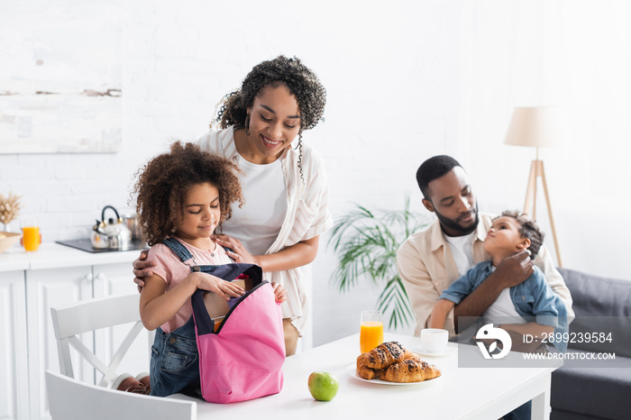 african american woman smiling near daughter packing backpack in kitchen