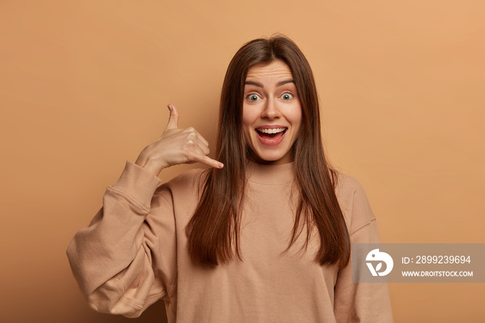 Communcation on distance concept. Happy European woman makes phone gesture, asks telephone number or give her call, smiles joyfully, dressed in casual sweatshirt, isolated on brown background.