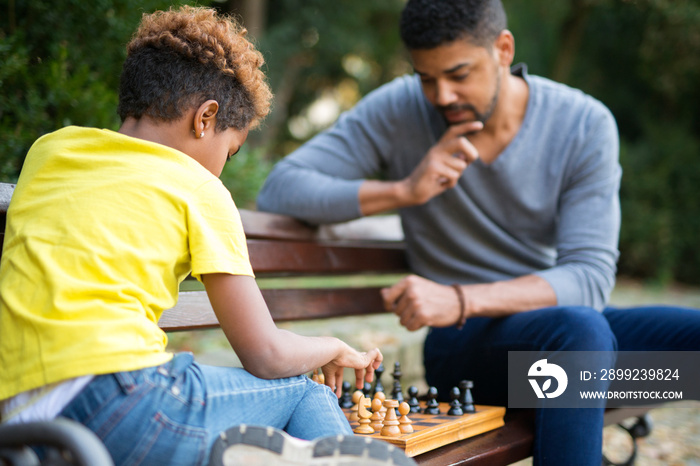 Father and daughter playing chess on the bench in city park. Family leisure time.