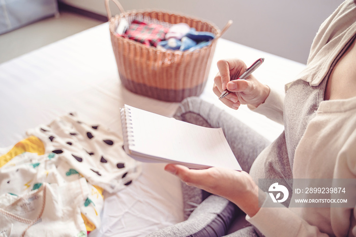 close up of pregnant woman writing on notebook sitting on bed with baby clothing in a basket and on the bed. resting and relaxing in the bedroom, wearing stretcher pants and cardigan with sun shine