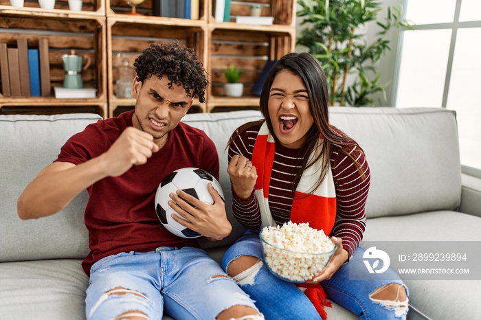 Young latin couple watching and supporting football match sitting on the sofa at home.