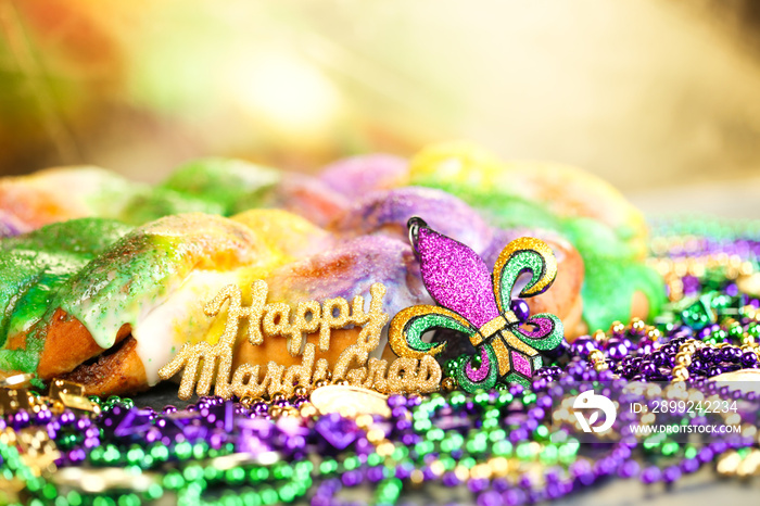 Happy Mardi Gras text in gold glitter and a king cake with yellow, green, and purple sprinkles surrounded by Mardi Gras beads and a glittering fleur de lis.