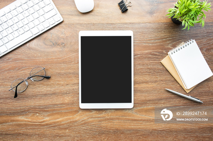 White tablet with black blank screen is on top of wood desk table with blank notebook and supplies. Top view, flat lay.