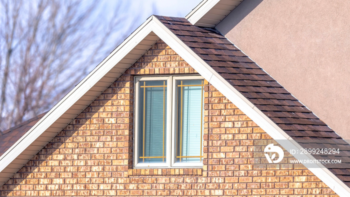 Panorama Close up of a home exterior with sunlit pitched roof over window and brick wall