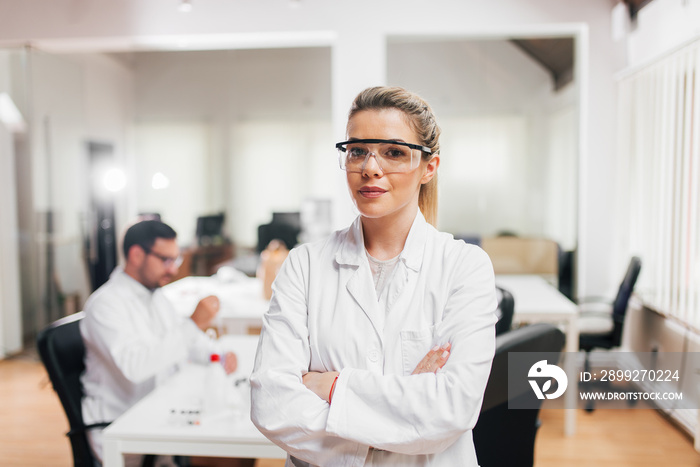 Portrait of a female scientist in protective eye wear with crossed arms. Colleagues in the background.