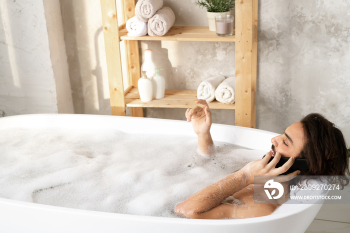 Young restful man talking by smartphone while enjoying hot bath with foam