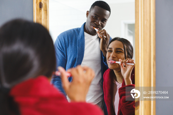 Happy diverse couple in brushing teeth in bathroom, looking in mirror smiling