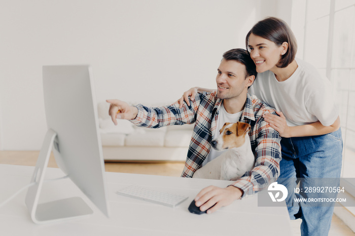 Affectionate brunette wife supports husband with doing project work, happy man points into monitor, tells something, shares his ideas, works together with pedigree dog, pose in spacious cabinet