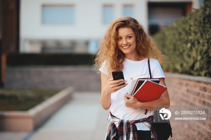 Female student using smart phone and holding books while walking in campus.