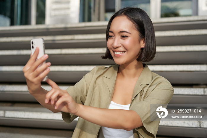 Portrait of asian beautiful girl takes selfie on smartphone. Young korean woman sits on stairs outdoors and makes photos on mobile phone