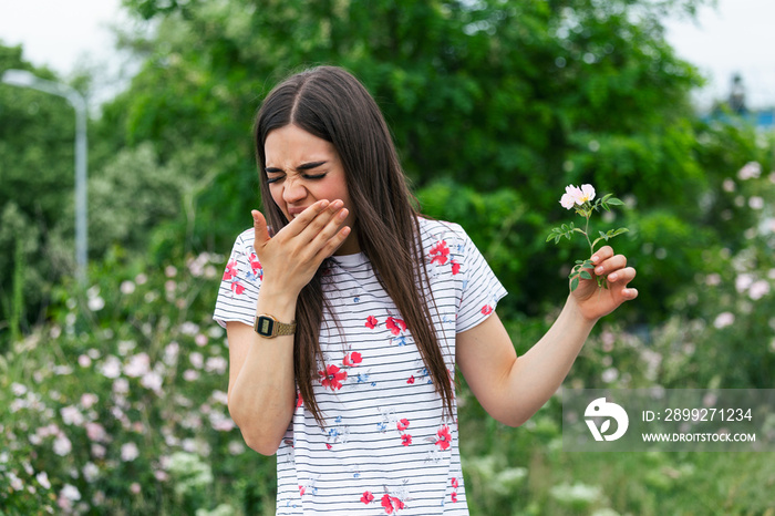 Young woman with Pollen allergy holding a flower and saying no.. Young woman with pollen and grass allergies. Flowering trees in background. Spring Seasonal allergies and health problems.