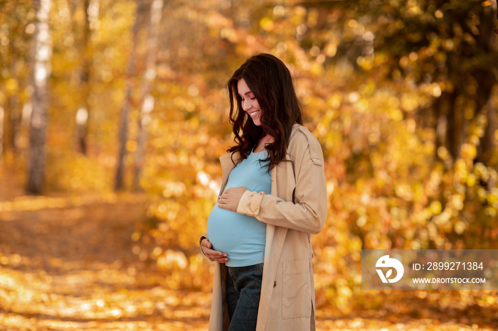 Gorgeous pregnant woman hugging her belly while walking at autumn park on beautiful sunny day