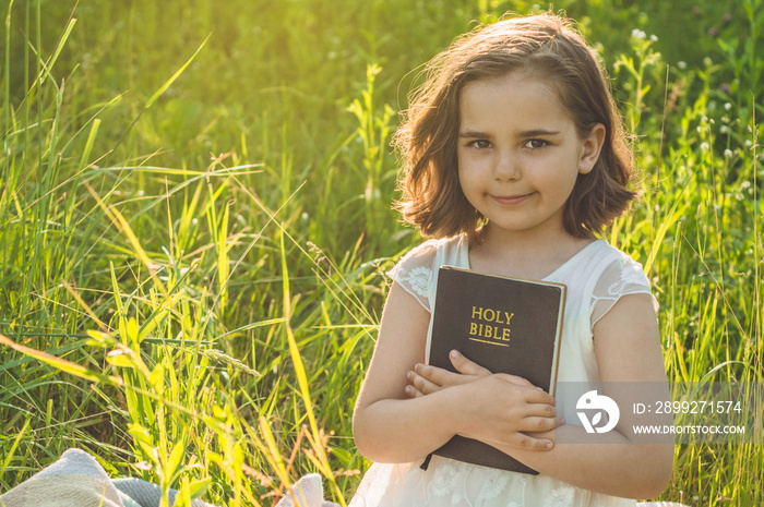 Christian girl holds bible in her hands. Reading the Holy Bible in a field during beautiful sunset. Concept for faith, spirituality and religion