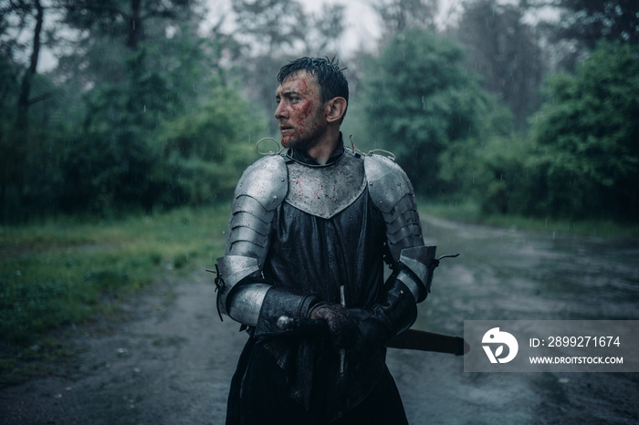 A young man stands in the image of a medieval knight in armor and with a sword.