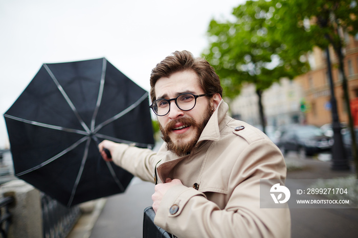 Bearded man in eyeglasses and trench-coat holding his umbrella in stormy weather