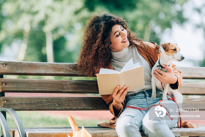 laughing woman cuddling jack russell terrier dog while sitting on bench with book