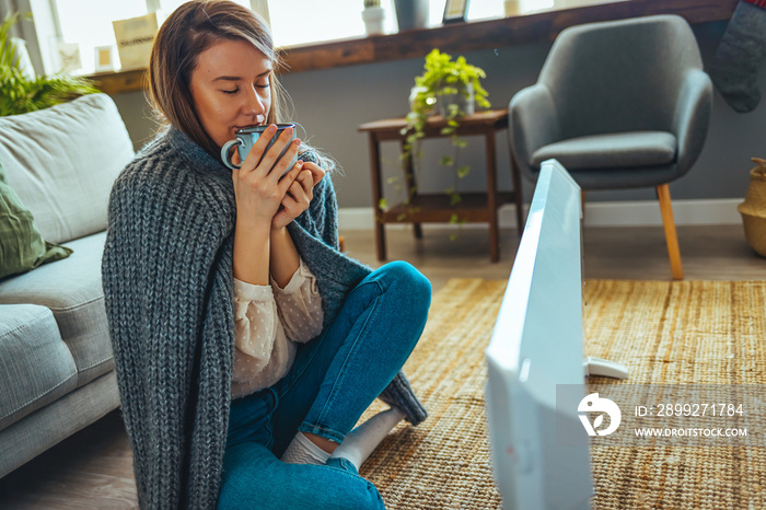 Girl squatting beside radiator with cup of hot tea in hands and trying to warm up. Grl in jacket holding hot tea beside radiator. Young woman sitting in room with electric heater.