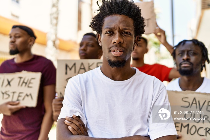 Young activist man with arms crossed gesture standing with a group of protesters holding banner protesting at the city.